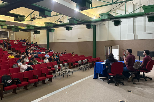 Imagem: A imagem mostra um auditório com poltronas vermelhas ocupadas por um público disperso, assistindo a um painel de palestrantes sentados em uma mesa coberta com uma toalha azul. O ambiente possui teto alto com estrutura metálica verde e iluminação distribuída. Os palestrantes, três homens e uma mulher, estão sentados em cadeiras giratórias e parecem estar discutindo um tema, com um dos homens gesticulando enquanto fala. O público, composto por homens e mulheres, está atento, com alguns fazendo anotações ou segurando objetos pessoais. No fundo, há uma porta branca e um extintor de incêndio fixado na parede.