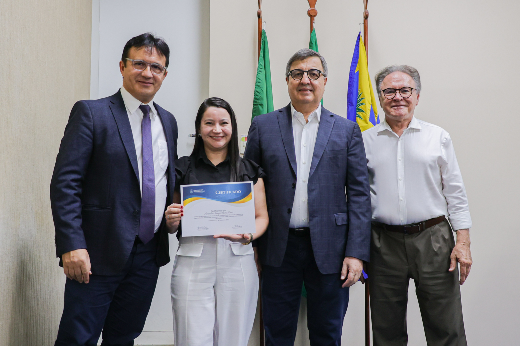 Imagem: quatro pessoas posando para uma foto em um ambiente institucional. Ao centro, uma mulher de cabelos escuros e lisos, vestindo blusa preta e calça branca, segura um certificado com as duas mãos e sorri. Ao seu redor, três homens vestindo roupas formais também sorriem. O homem à esquerda usa terno azul-escuro e gravata roxa, enquanto o homem ao centro usa um blazer azul e camisa branca. O homem à direita veste uma camisa branca e calça marrom. Ao fundo, há três bandeiras, incluindo a do Brasil e de um estado brasileiro.