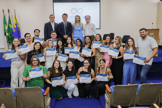 Imagem: grupo de aproximadamente 25 pessoas reunidas em um auditório para uma cerimônia de entrega de certificados. A maioria dos participantes são mulheres, segurando seus certificados e sorrindo para a câmera. Na parte de trás, há três homens e uma mulher de pé, vestidos com roupas formais, indicando serem organizadores ou autoridades do evento. O ambiente tem cadeiras acolchoadas, bandeiras do Brasil e de um estado brasileiro ao fundo, além de um telão exibindo um logotipo com o símbolo do infinito e a palavra "Conectea". O tapete azul e o púlpito sugerem um ambiente acadêmico ou institucional.