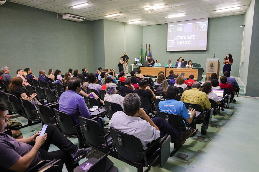 Imagem: A imagem mostra um auditório com paredes verdes, onde ocorre um evento da Universidade Federal do Ceará (UFC). No palco, há uma mesa com cinco pessoas sentadas, enquanto uma mulher em pé, à direita, faz gestos, possivelmente interpretando em Língua Brasileira de Sinais (Libras). No telão ao fundo, lê-se "ESTATUINTE DA UFC". O público, composto por diversas pessoas sentadas em cadeiras pretas, acompanha a apresentação. Algumas pessoas fazem anotações ou usam celulares. Bandeiras estão posicionadas ao lado do palco, e um fotógrafo registra o evento.