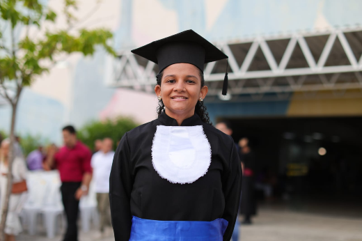 Imagem: Uma mulher sorri enquanto posa para a foto, vestindo beca de formatura preta com gola branca decorativa, faixa azul na cintura e capelo (chapéu de formatura) na cabeça. Ela está em pé ao ar livre, em frente a um prédio com estrutura metálica visível e algumas pessoas desfocadas ao fundo. Há uma árvore à esquerda e o clima parece agradável.