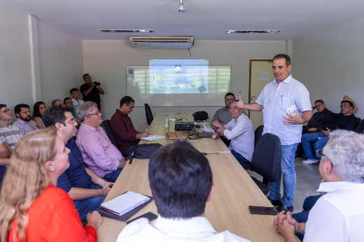 Imagem: Em uma sala de reunião clara e simples, cerca de 18 pessoas estão reunidas em torno de uma mesa retangular de madeira. Um homem em pé, à direita da imagem, fala para o grupo, gesticulando com uma das mãos. Ele veste camisa branca e calça jeans. Os demais participantes, homens e mulheres, estão sentados, atentos à apresentação, alguns com cadernos e laptops sobre a mesa. Ao fundo, um projetor exibe uma imagem com brasões e a palavra "Quixadá". Há também uma pessoa em pé ao fundo registrando o momento com uma câmera.