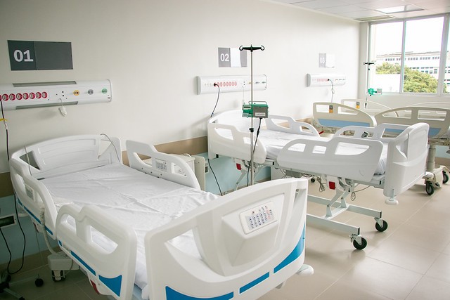 Imagem: A imagem mostra um quarto hospitalar moderno e bem iluminado. No ambiente, há três leitos hospitalares numerados (01, 02 e 03), todos equipados com camas ajustáveis eletronicamente, com controles embutidos nas laterais das camas. Cada leito possui também um painel na parede com botões de chamada de enfermagem e conexões para gases medicinais (oxigênio, ar comprimido, etc.), além de suportes para equipamentos intravenosos. O ambiente é limpo, organizado e claro, com iluminação natural vinda de uma janela ampla ao fundo, o que contribui para uma atmosfera mais acolhedora e confortável para os pacientes.