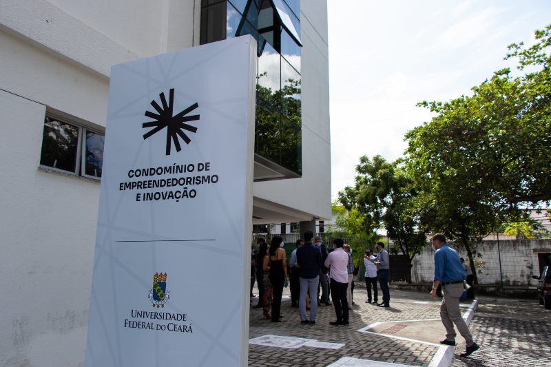 Imagem: A imagem mostra a entrada de um prédio moderno com uma placa grande em destaque. Na placa, lê-se:  "CONDOMÍNIO DE EMPREENDEDORISMO E INOVAÇÃO" "UNIVERSIDADE FEDERAL DO CEARÁ"  Abaixo do texto está o brasão da universidade.  Na frente do prédio, há várias pessoas reunidas, possivelmente participando de um evento. O ambiente é urbano, com árvores ao redor e piso de blocos intertravados. A fachada do edifício é branca, com janelas espelhadas, refletindo a vegetação ao redor. A cena ocorre durante o dia, sob luz natural.