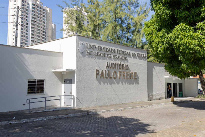 Imagem: A imagem mostra a fachada do Auditório Paulo Freire, localizado na Faculdade de Educação da Universidade Federal do Ceará (UFC). A construção é de cor clara, com letras metálicas em relevo destacando o nome da instituição e do auditório. À esquerda, há uma rampa de acessibilidade ao lado de uma pequena janela. À direita, há uma entrada maior com portas de vidro, parcialmente abertas, por onde se vê uma pessoa no interior.  Ao fundo, vemos edifícios residenciais altos, árvores e céu claro, sugerindo que o local está situado em uma área urbana bem arborizada. Há também uma grande árvore com sombra à direita, que contrasta com o cimento do pátio frontal. O ambiente é limpo e bem conservado.