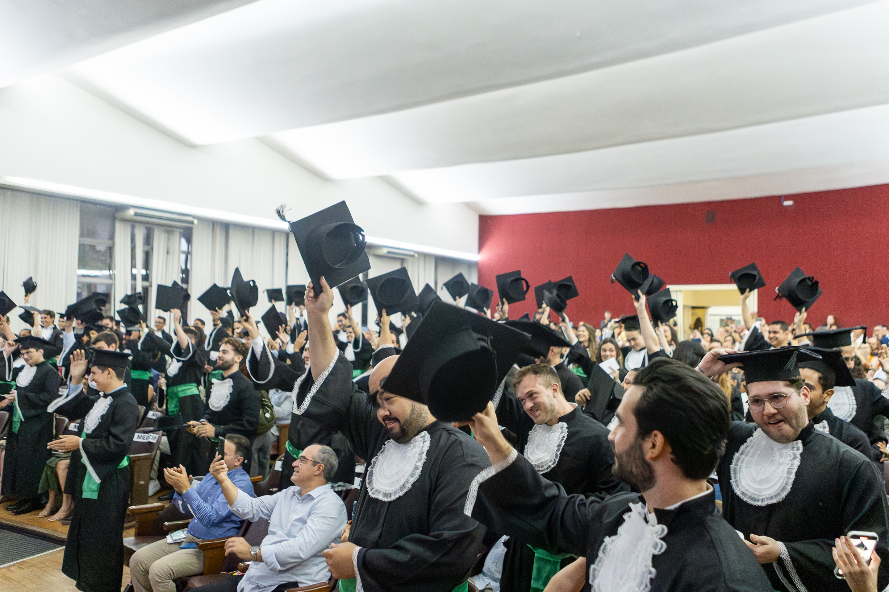 Imagem: grupo de formandos vestidos com becas pretas com detalhes brancos e faixas verdes celebra em pé dentro de um auditório, erguendo os capelos (chapéus de formatura) no ar com entusiasmo. O ambiente está lotado, com familiares e amigos sentados nas cadeiras ao redor, acompanhando a cerimônia. O clima é de alegria e comemoração.