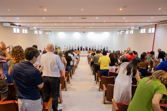 Imagem: foto tirada do fundo de um auditório lotado, onde o público, em pé entre fileiras de cadeiras vermelhas, aplaude os integrantes da mesa de honra posicionados ao fundo do palco. Ao todo, cerca de vinte pessoas estão sentadas à mesa, voltadas para a plateia. Atrás delas, à esquerda, estão três bandeiras: do Brasil, do estado do Ceará e da Universidade Federal do Ceará (UFC). O ambiente é amplo, com paredes claras e iluminação de teto.
