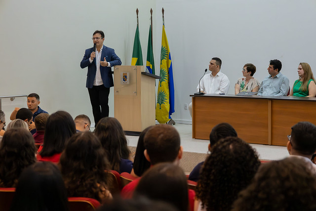 Imagem: em auditório com fileiras de cadeiras vermelhas ocupadas por estudantes, um homem de terno azul e camisa branca fala ao microfone em pé, próximo a um púlpito com o brasão da Universidade Federal do Ceará (UFC). Atrás dele, estão hasteadas três bandeiras: do Brasil, do estado do Ceará e da UFC. À direita da imagem, uma mesa com cinco pessoas sentadas, entre homens e mulheres, acompanha a fala. Todos os presentes observam atentamente.