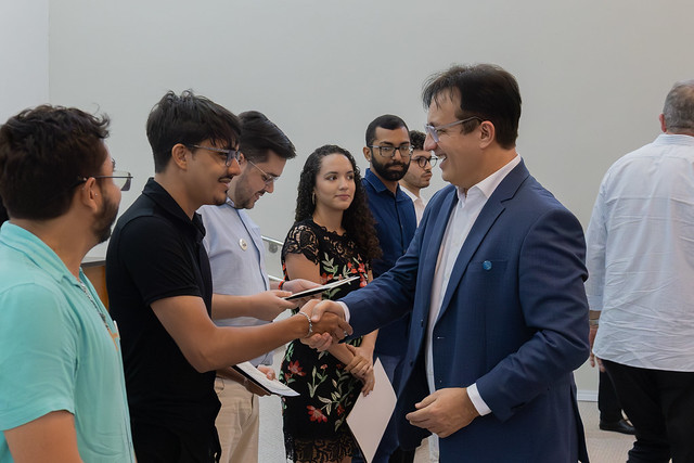 Imagem: em ambiente interno, um homem de terno azul e camisa branca, usando óculos, sorri enquanto aperta a mão de um jovem de camiseta preta e óculos, que segura um certificado. Ao redor deles, outras pessoas observam a interação: um rapaz de camiseta azul clara, outro de camisa branca, uma jovem de vestido preto com estampa floral e dois homens de camisa social. Ao fundo, há uma parede clara e pessoas de costas.