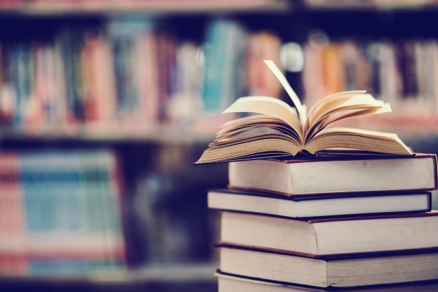Imagem: A imagem mostra uma pilha de livros em primeiro plano, com um livro aberto no topo. As páginas do livro aberto estão levemente curvadas, sugerindo movimento ou que foi recém-folheado. Ao fundo, desfocado, é possível ver várias estantes cheias de livros, criando o ambiente típico de uma biblioteca ou livraria. As cores são suaves, com tons quentes que passam uma sensação de tranquilidade e aconchego, reforçando a ideia de estudo, leitura ou pesquisa