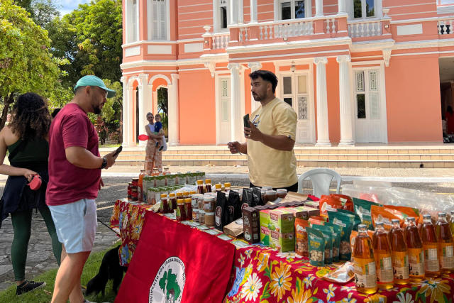 Imagem: A imagem mostra uma feira ao ar livre realizada no jardim da Reitoria da Universidade Federal do Ceará (UFC), em frente ao prédio histórico de fachada cor-de-rosa com detalhes brancos. Em primeiro plano, há uma barraca coberta com uma toalha colorida estampada e uma bandeira do Movimento dos Trabalhadores Rurais Sem Terra (MST). Sobre a mesa estão expostos diversos produtos agroecológicos, como mel, cafés, biscoitos e outros itens alimentícios.  Um expositor, vestindo camisa bege, conversa com um visitante que usa boné azul, camiseta bordô e bermuda clara. Outras pessoas circulam ao fundo, incluindo uma mulher com roupas esportivas segurando uma garrafa e um homem que segura uma criança no colo. O clima é de dia ensolarado, e o evento parece valorizar a agricultura familiar e a integração entre universidade e comunidade.