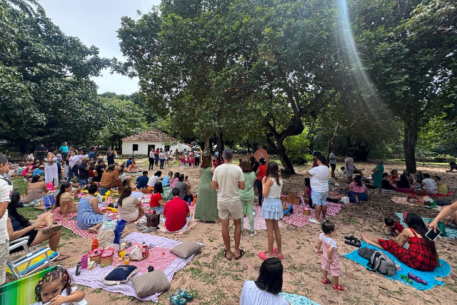 Imagem: A imagem mostra um grande piquenique coletivo nos jardins da Casa José de Alencar, em Fortaleza. Famílias e crianças estão sentadas ou deitadas em toalhas coloridas e mantas estendidas sobre o chão de terra batida, sob a sombra de árvores frondosas. Alguns adultos estão em pé, acompanhando uma apresentação que ocorre mais ao fundo, próxima à casa histórica de fachada branca e telhado de barro.  O clima é de descontração e convivência ao ar livre, com crianças brincando e pessoas conversando. Detalhes como almofadas, lanches, garrafas e brinquedos reforçam o ambiente acolhedor e familiar, característico de um evento cultural que integra lazer, patrimônio histórico e encontro comunitário