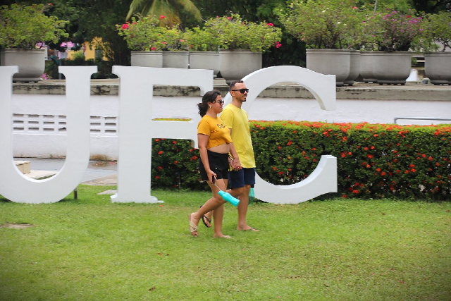 Imagem: A imagem mostra um casal caminhando pelos jardins da Reitoria da Universidade Federal do Ceará (UFC), durante as atividades do projeto “UFC de Portas Abertas”. Eles passam em frente a grandes letras brancas que formam a sigla “UFC”. Ao fundo, há um canteiro florido com arbustos de flores vermelhas e grandes vasos de plantas ornamentais sobre um muro. O homem veste uma camisa amarela e shorts escuros, enquanto a mulher usa uma blusa amarela, shorts pretos e carrega uma garrafa azul. O clima parece agradável, reforçando o ambiente acolhedor e descontraído do evento