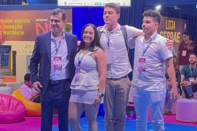 Imagem: quatro pessoas estão em pé sobre um palco azul iluminado, posando para foto. À esquerda, está um homem de terno azul-marinho, camisa clara e crachá pendurado no pescoço. Ao seu lado, uma mulher veste roupa branca sem mangas e sandálias. Os dois homens seguintes também usam roupas claras, com crachás, e um deles carrega uma mochila. Ao fundo, há placas com as palavras “Loja Sebrae” e “Sebrae NE”, além de um telão com os dizeres “Ideathon da Saúde HU e UFPI — R$ 3.000, 2º colocado”. O ambiente tem iluminação colorida e clima festivo.