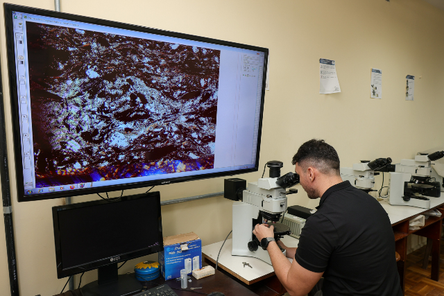 Imagem: Em uma sala de laboratório, um homem de pele clara, cabelo escuro e curto, veste camisa preta e está sentado, olhando através de um microscópio. À frente dele, um monitor grande exibe uma imagem ampliada com padrões coloridos em tons escuros, brancos, azulados e avermelhados, possivelmente de uma amostra mineral ou geológica. Ao lado do microscópio, há uma mesa com um computador, uma caixa de luvas descartáveis, tubos de ensaio e outros materiais de laboratório. Ao fundo, observa-se uma fileira de outros microscópios sobre bancadas e alguns cartazes fixados na parede clara