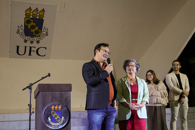 Imagem:  Em um palco iluminado, um homem de óculos, vestindo blazer preto, camisa laranja escura e calça jeans, fala ao microfone sorrindo. Ao lado dele, uma mulher de cabelos grisalhos, com blazer verde-claro e roupa vermelha, o observa com expressão simpática. Atrás deles, outras três pessoas acompanham a fala: duas mulheres — uma de blusa bege e óculos — e um homem de blazer claro e calça preta. À esquerda da imagem, há um púlpito de madeira com o brasão da Universidade Federal do Ceará (UFC), e, ao fundo, uma parede clara exibe em destaque o mesmo brasão e a sigla “UFC”.