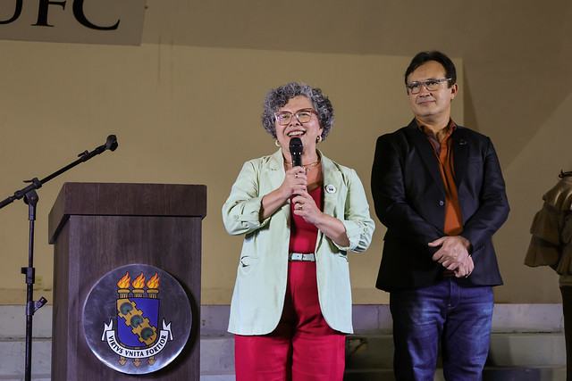 Imagem: Em um palco iluminado, uma mulher de cabelos grisalhos e encaracolados, usando óculos, um blazer verde-claro e roupa vermelha, fala ao microfone com um sorriso no rosto. Ela está posicionada ao lado de um púlpito de madeira com o brasão da Universidade Federal do Ceará (UFC). Atrás dela, um homem de óculos, com camisa laranja escura e blazer preto, a observa atentamente com as mãos cruzadas. Ambos estão em pé sobre degraus claros, e ao fundo vê-se parte da palavra “UFC” em uma faixa branca.