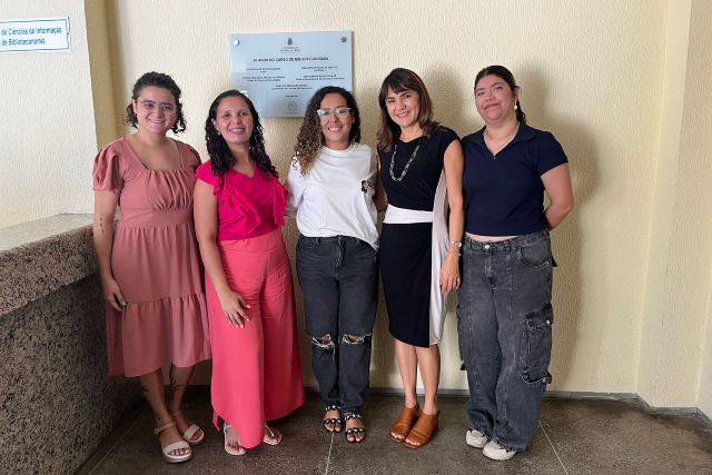 Imagem: Cinco mulheres posam sorrindo para a foto em um ambiente interno, de frente para uma parede clara com uma placa comemorativa metálica fixada. À esquerda, há uma bancada de pedra. Da esquerda para a direita: a primeira mulher usa vestido rosa claro e sandálias; a segunda, blusa rosa vibrante e calça pantalona no mesmo tom; a terceira, camiseta branca e calça jeans rasgada nos joelhos; a quarta, vestido preto com faixa branca na cintura e sandálias marrons; e a quinta, blusa preta e calça jeans larga com bolsos. Todas estão em pé, próximas umas das outras.