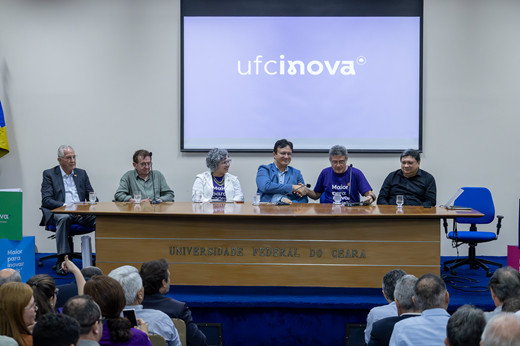 Imagem: Registro da mesa solene do evento de lançamento da Agência de Inovação UFC Inova mostra  as autoridades sentadas na frente do telão; o público contempla de costas