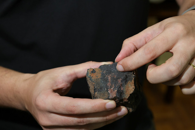 Imagem: duas mãos seguram um pedaço irregular de rocha escura, com manchas avermelhadas e aspecto poroso. Um dos dedos da pessoa aponta para a superfície da pedra, destacando sua textura e detalhes minerais.