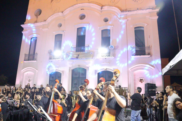 Imagem: Apresentação de uma orquestra em espaço aberto, à noite, em frente a um prédio histórico iluminado com luzes coloridas. Na primeira fila, músicos tocam contrabaixos, vestidos de preto. Atrás deles, outros instrumentistas e um coral acompanham a execução. O público observa a apresentação em pé, em clima de concerto festivo.
