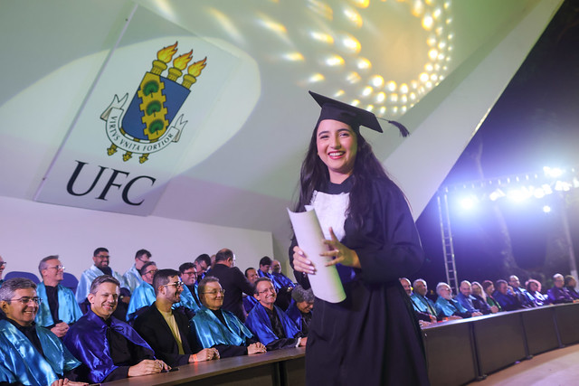 Imagem: jovem formanda com beca preta e capelo sorri segurando o diploma enrolado, em frente à mesa de honra da cerimônia. Atrás dela, professores com capas azuis e roxas estão sentados. No alto, aparece o brasão da Universidade Federal do Ceará (UFC) projetado na parede do palco iluminado.