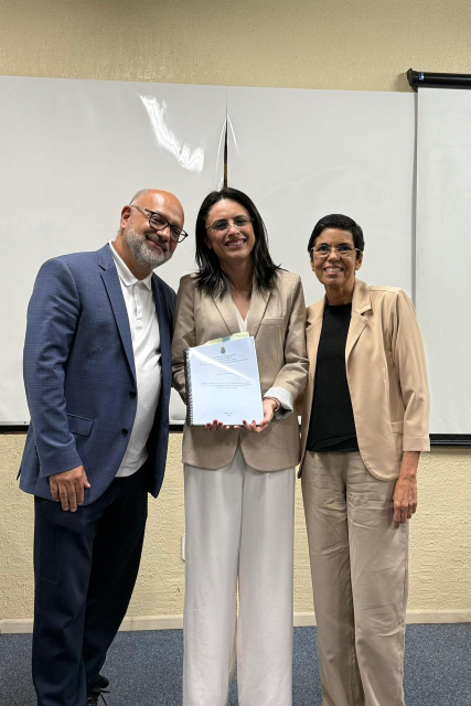 Imagem: Três pessoas estão lado a lado, sorrindo para a câmera. No centro, uma mulher de cabelos pretos lisos, usando óculos, blazer bege e calça clara, segura um volume encadernado com capa branca, semelhante a uma dissertação ou tese. À esquerda, um homem careca, de barba grisalha, óculos, camisa branca e blazer azul. À direita, uma mulher de cabelos curtos e óculos, vestindo blazer e calça bege com blusa preta. Ao fundo, há um quadro branco fixado na parede.