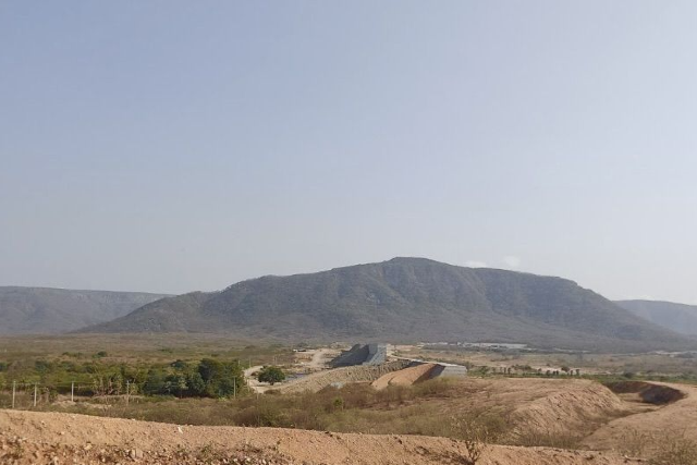 Imagem: #ParaTodosVerem: Paisagem com uma serra ao fundo, sob céu claro e azul. Em primeiro plano, aparece um terreno de solo avermelhado e seco, típico do semiárido, com áreas de vegetação esparsa. No centro da imagem, é possível ver uma grande estrutura de concreto que se estende em linha reta, lembrando um canal ou obra de engenharia, cortando a paisagem em direção à serra. Há também pequenas áreas verdes com árvores e construções espalhadas pela planície.