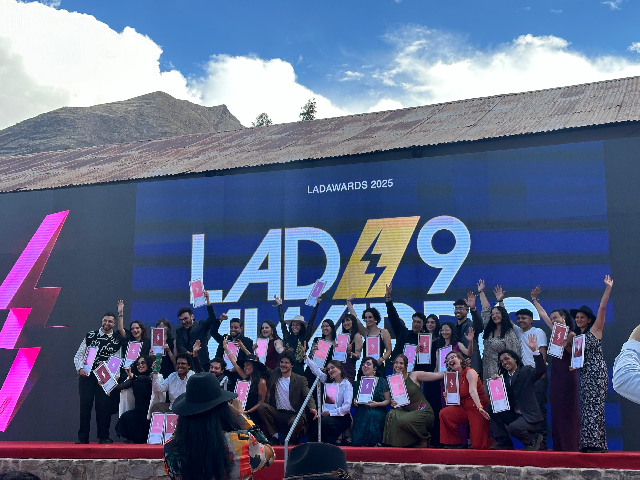 Imagem: Foto posada de um grupo de 27 pessoas em um palco vermelho, segurando placas de premiação. A maioria está com as mãos levantadas. Atrás deles há uma grande placa azul, onde se lê Lad 9 Awards (Foto: Divulgação)