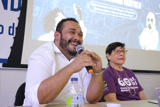 Imagem:Um homem de barba, com camisa branca, sorri enquanto fala ao microfone. Ao seu lado está uma mulher de óculos e cabelos curtos, também de camiseta roxa dos 60 anos do curso de Engenharia Química da UFC. Ambos participam da mesa-redonda no evento comemorativo.