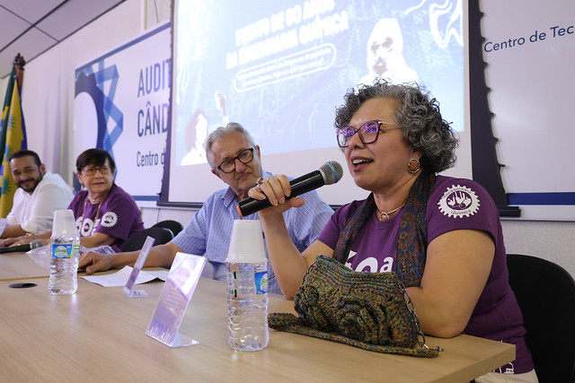 Imagem: Uma mulher de óculos e cabelos cacheados grisalhos fala ao microfone, sorridente. Ela veste uma camiseta roxa com o logo dos 60 anos do curso de Engenharia Química da UFC. Ao lado dela, outros três integrantes da mesa-redonda acompanham atentos.