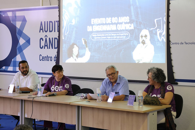 Imagem: Quatro pessoas estão sentadas atrás de uma mesa em um auditório, participando de uma mesa-redonda. Duas mulheres usam camisetas roxas com o logo dos 60 anos do curso de Engenharia Química da UFC, enquanto um homem de camisa azul clara e outro de camisa branca também compõem a mesa. Ao fundo, um telão exibe a mensagem “Evento de 60 anos da Engenharia Química”.