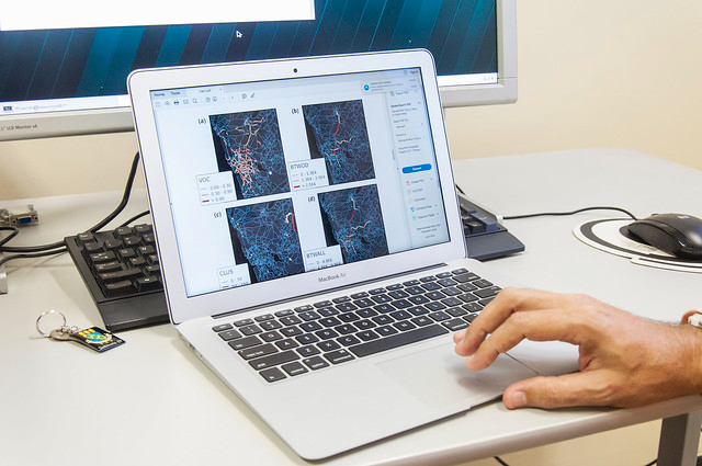 Imagem: Fotografia mostra uma mão masculina sobre o teclado de um notebook prateado. Na tela do computador portátil, aparecem gráficos científicos em forma de imagens coloridas com linhas e pontos interligados, acompanhados de legendas e siglas. O notebook está sobre uma mesa clara, ao lado de um teclado preto, um mouse e um chaveiro com crachá. Ao fundo, vê-se parte de um monitor maior.