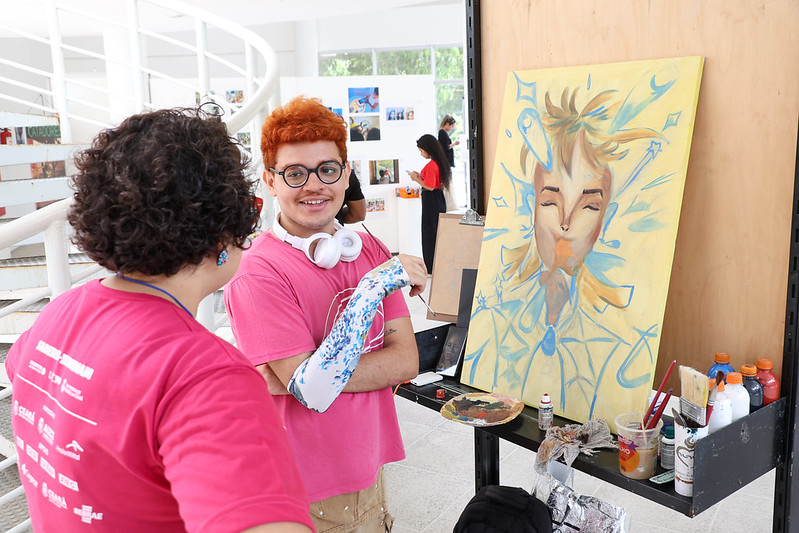 Imagem: Na imagem, duas pessoas estão em um ambiente interno claro e moderno, durante a Mostra ICA 2024. Ambas vestem camisetas rosas.  Em primeiro plano, uma delas — de cabelo curto e ruivo, óculos redondos e fones brancos pendurados no pescoço — está sorrindo enquanto segura pincéis. À sua frente, há uma tela apoiada em um painel, com uma pintura colorida de um rosto estilizado, em tons de amarelo e azul, cercado por formas geométricas e abstratas que lembram respingos e estrelas.  Na lateral da tela, há pincéis, tintas e recipientes de apoio. Ao fundo, outras pessoas observam imagens expostas na parede branca, compondo o cenário do evento artístico coletivo.