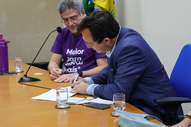 Imagem: dois homens estão sentados à mesa em um auditório. Um deles, de terno azul, assina um documento. Ao lado, outro homem, de camiseta roxa, observa.