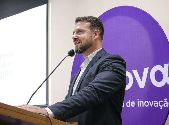 Imagem: Homem de barba curta e cabelo castanho fala ao microfone em um púlpito de madeira. Ele veste terno escuro e camisa branca. Ao fundo, há um grande painel circular roxo com o logotipo da Agência UFC Inova.