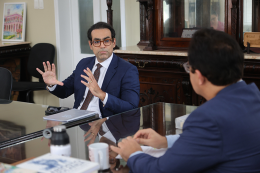 Imagem: Em primeiro plano, o reitor Custódio Almeida de costas; de frente para a câmera, o juiz Júlio Rodrigues gesticula. Ambos estão sentados e vestem ternos azuis.