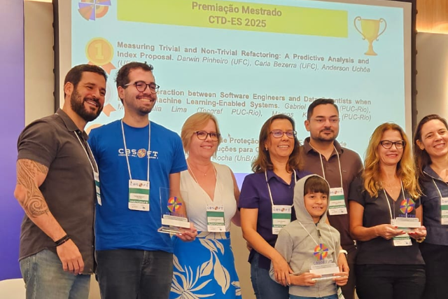 Imagem: Grupo de oito pessoas posando sorridentes para foto em ambiente de evento acadêmico. Três delas seguram troféus de premiação com o logo do CBSoft. Ao centro, uma criança também segura um troféu e está vestida com um moletom cinza com capuz. Ao fundo, há uma projeção em tela com o título "Premiação Mestrado – CTD-ES 2025", seguido dos nomes dos trabalhos premiados e autores, com destaque para o trabalho "Measuring Trivial and Non-Trivial Refactoring", de Darwin Pinheiro, Carla Bezerra e Anderson Uchôa, da UFC. Todos estão usando crachás do evento.