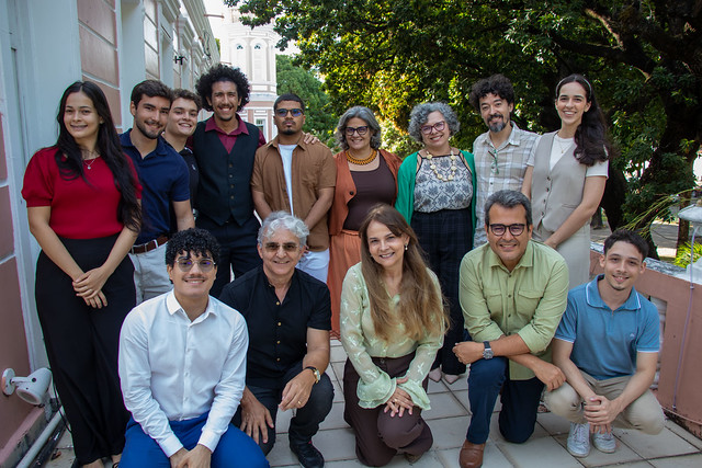 Imagem: Grupo de treze pessoas posa sorridente em área externa, diante de prédio histórico de cor rosa e branco, com árvores ao fundo. Há homens e mulheres de idades diversas, alguns agachados na frente e outros em pé atrás. Todos estão vestidos de forma social ou casual arrumada, formando uma composição descontraída e acolhedora para a fotografia.