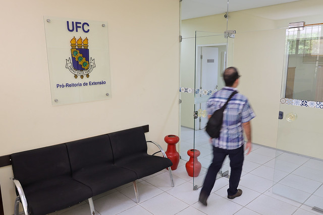 A imagem mostra o interior de um prédio, especificamente da Pró-Reitoria de Extensão (PREX) da Universidade Federal do Ceará (UFC). No lado esquerdo da imagem, há uma placa na parede com o brasão da UFC e os dizeres "Pró-Reitoria de Extensão".  No centro da imagem, um homem de costas está caminhando em direção a uma porta de vidro, que dá acesso a outra sala ou corredor. A sala tem piso de cerâmica branca e paredes claras, transmitindo um ambiente limpo e organizado. O local parece ser uma área de recepção ou espera dentro da pró-reitoria. 