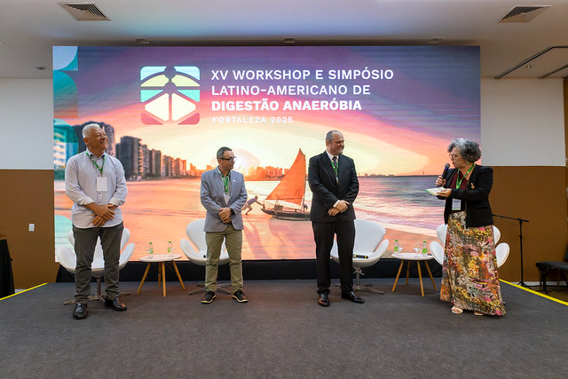 Na manhã do dia 14, a professora Diana Azevedo, vice-reitora da UFC, moderou a mesa sobre o papel da digestão anaeróbia na transição energética e descarbonização (Foto: Guilherme Silva/UFC Informa) Imagem: Em um auditório, quatro pessoas estão em pé no palco diante de um grande painel digital com a identidade visual do XV Workshop e Simpósio Latino-Americano de Digestão Anaeróbia – Fortaleza 2025. Ao fundo, o painel exibe a imagem de uma praia com jangada ao entardecer. À direita, uma mulher de cabelos grisalhos fala ao microfone; à esquerda, três homens escutam atentamente. Cadeiras brancas e garrafas de água compõem o cenário.