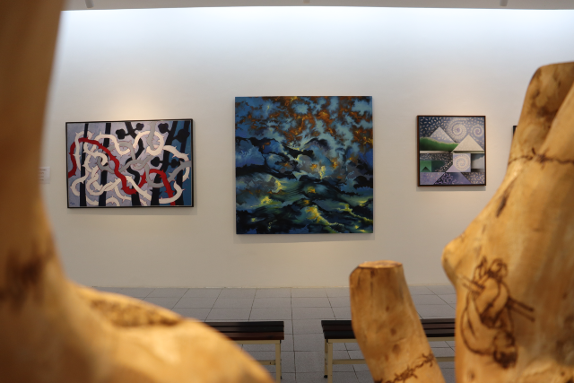 Na exposição "Acervo Mauc – Aquisições Recentes: 2ª edição", desenhos, gravuras, fotografias, esculturas, pinturas de artistas e colecionadores que doaram suas obras ao Mauc estão em exposição (Foto: Kathleen Silveira) Imagem: Sala de exposição com três pinturas penduradas em uma parede branca. As obras apresentam estilos e cores diferentes — uma com formas abstratas e linhas vermelhas, outra com tons de azul e dourado lembrando nuvens, e a terceira com figuras geométricas. Em primeiro plano, aparecem partes de esculturas em madeira com desenhos gravados, desfocadas, compondo a cena. No centro da sala, há dois bancos para os visitantes.