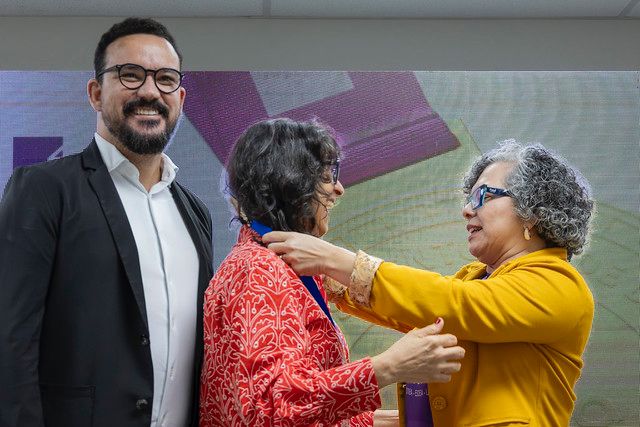 Homenageada pelo professor Carlos Adriano Santos Gomes Gordiano, a professora Mônica Cavalcanti Sá de Abreu recebe sua medalha das mãos da vice-reitora Diana Azevedo (Foto: Guilherme Silva/UFC). Na imagem, três pessoas estão em um momento de cerimônia. Ao centro, uma mulher de cabelos curtos e cacheados, usando roupa vermelha, recebe uma medalha das mãos de outra mulher, que veste um casaco amarelo e óculos. À esquerda, um homem de barba, óculos e terno escuro sorri enquanto acompanha a homenagem.