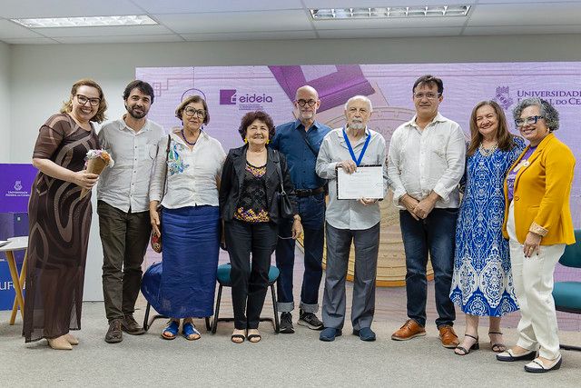 O Mentores da Docência é uma homenagem da UFC que reconhece professores que se destacam por suas contribuições à educação. Na foto, o professor Roberto Martins Castelo, do Instituto de Arquitetura, Urbanismo e Design, recebe a homenagem com um dos mentores da docência desta edição (Foto: Guilherme Silva/UFC) A foto mostra um grupo de oito pessoas posando lado a lado em um ambiente interno, durante uma cerimônia. Ao centro, um homem mais velho segura um certificado e usa uma medalha azul no pescoço, sugerindo que ele foi homenageado. As demais pessoas, homens e mulheres, sorriem e estão em trajes sociais, demonstrando clima de celebração e respeito. Ao fundo, há um painel com as logomarcas da Universidade Federal do Ceará (UFC) e da Eideia, indicando que o evento está ligado à instituição.