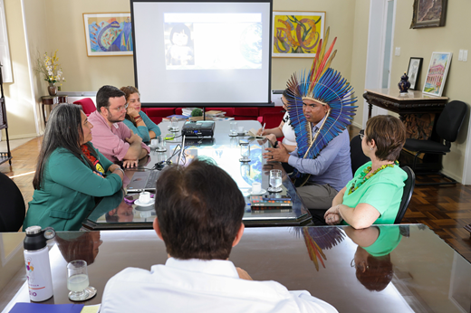 Reitor Custódio Almeida (de camisa azul, sentado de costas) reúne-se na mesa com os demais participantes da reunião (dois homens e quatro mulheres). Ao fundo, uma apresentação é projetada no telão do Gabinete da Reitoria.