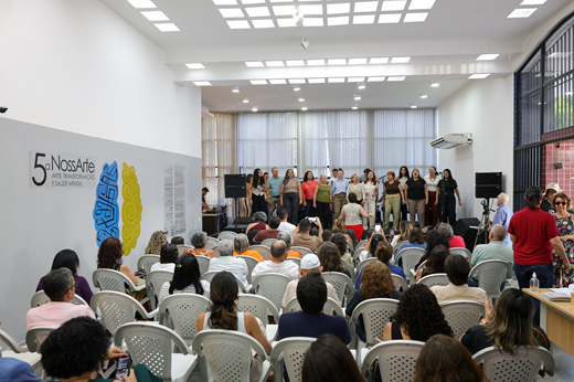 No dia 14 de outubro, o Mauc recebeu a abertura da exposição NossaArte, que reúne obras de servidores ativos, aposentados e trabalhadores terceirizados; mostra segue aberta à visitação até 14 de novembro. (Foto: Viktor Braga/ UFC Informa) Imagem: Sentado, público presente à abertura da Exposição NossaArte assiste à apresentação de coral dos servidores; à esquerda, vê-se um adesivo com a logomarca da exposição.