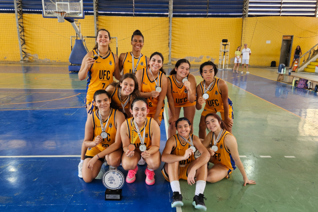 Na modalidade feminina, o campeonato foi decidido entre a Universidade Federal do Pará (UFPA) e a UFC. A equipe cearense ganhou medalha de prata (Foto: Divulgação/Secretaria de Esportes da UFC) Imagem: Grupo de dez atletas do time feminino de basquete da Universidade Federal do Ceará (UFC) posa para foto em quadra esportiva coberta, com piso em tons de azul e verde e paredes amarelas ao fundo. As jogadoras, que vestem uniformes amarelos com detalhes azuis e a sigla “UFC” estampada no peito, estão sorrindo e exibem medalhas penduradas no pescoço. À frente do grupo, no chão, há um troféu preto e prateado. Ao fundo, vê-se uma tabela de basquete e parte da arquibancada.