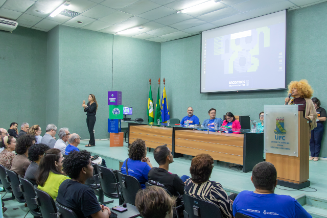 Imagem: foto do audiório reitor Ícaro Moreira, mostra aplateia de costas, sentada em cadeiras, e o palco do evento, com uma mesa onde estão sentados os participantes da solenidade e, no ´pulpito, a estudante Angela Abuk