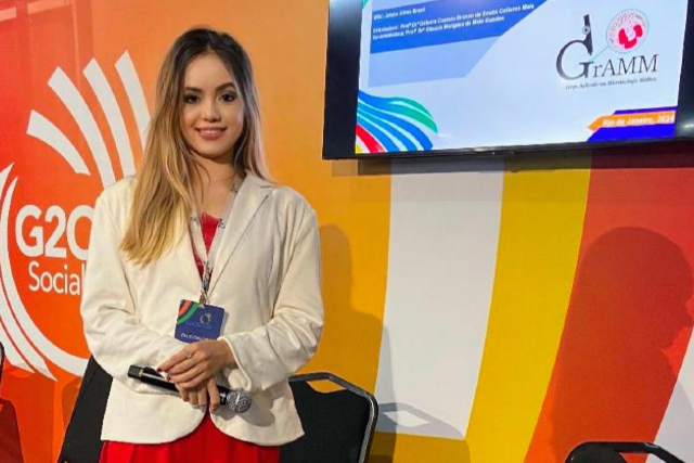 Imagem: uma mulher está em pé em um evento, com um microfone na mão. Ela veste blazer claro e vestido vermelho, e atrás dela há um painel colorido com o logotipo “G20 Social”
