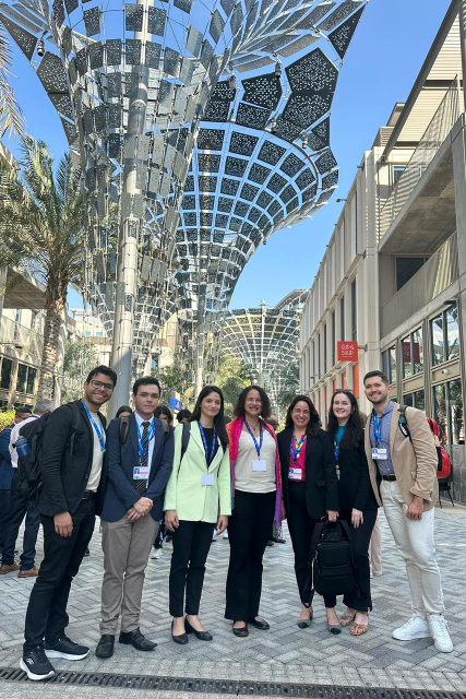 Imagem: sete pessoas posam sorrindo em um ambiente moderno e ensolarado, com estruturas metálicas no alto que refletem a luz. Todas usam crachás e roupas formais, sugerindo participação em um evento internacional